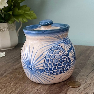 May include: A white ceramic jar with a blue lid and a blue and white pine cone design. The jar has a rustic, handmade look.