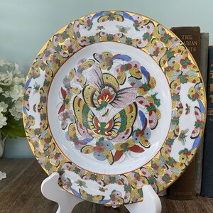 May include: A white porcelain plate with a gold rim and a colourful butterfly design. The butterflies are painted in shades of blue, red, yellow, and green. The plate is on a white stand.