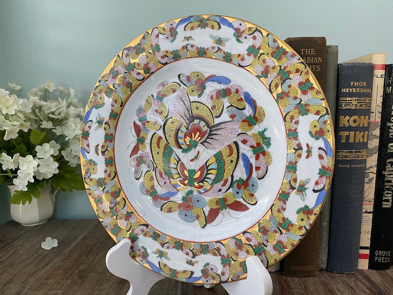 May include: A white porcelain plate with a gold rim and a colourful butterfly design. The butterflies are painted in shades of blue, red, yellow, and green. The plate is on a white stand.