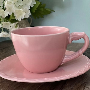 May include: A pink teacup and saucer set. The cup has a unique handle design and sits on a matching saucer with a decorative edge. The set is made of ceramic and is a soft pink color.