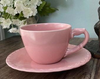 May include: A pink teacup and saucer set. The cup has a unique handle design and sits on a matching saucer with a decorative edge. The set is made of ceramic and is a soft pink color.