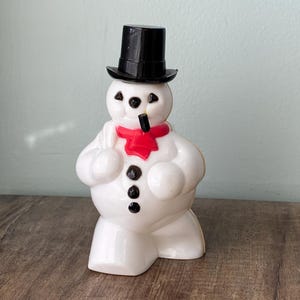 May include: A white snowman figurine with a black top hat, black eyes, and a black mouth. It has a red scarf and black buttons. The snowman is a decorative item.