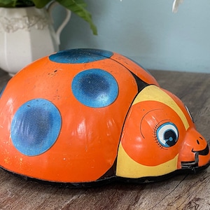 May include: A vintage wind-up toy ladybug with orange and yellow paint, blue spots, and a black outline. The toy has a smiling face with large blue eyes and eyelashes.