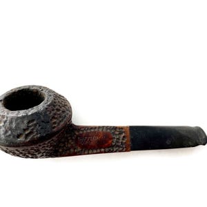 May include: A tobacco pipe with a textured, dark brown bowl and a black stem. The bowl has a rough, pitted surface. The stem is connected to a brown section with text. The pipe is set against a white background.