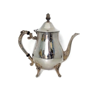 May include: A silver-toned metal teapot featuring a polished, reflective body, a curved spout, and an ornate handle with two black accents. The teapot stands on three decorative feet and includes a lid topped with a small, sculpted finial.
