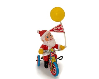 May include: A red and white wind-up toy Santa Claus riding a red, blue, and yellow tricycle. Santa is wearing a white beard and a red hat. He is holding a yellow balloon with a red flag that says "Merry Christmas!"