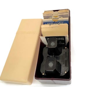 May include: A vintage View-Master set with a beige case and a burgundy box. The box contains a black viewer, instruction sheets, and several View-Master reels. The case is open, revealing the contents.