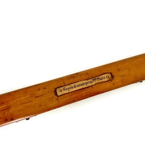 May include: A long, rectangular wooden box with a light brown finish. A small, rectangular label with the text "Regolo di convergenza MP Maltei" is affixed to the top. The box is angled, with a white background.