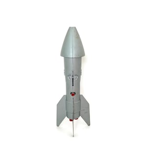 May include: A silver-colored toy rocket ship featuring a pointed nose cone, three large stabilizing fins, and a vertical mechanical lever on the side. The rocket includes a small red circular detail and red circular components at the base.