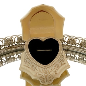 May include: Ivory-colored heart-shaped ring box with a black velvet interior. The box has ornate floral detailing and is open, revealing a ring. A silver-toned decorative tray with floral accents frames the box.