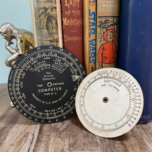 May include: Two vintage circular aviation slide rules. One is black with white markings, labeled "COMPUTER TYPE D-4" and "PROPERTY OF U.S. ARMY AIR FORCES." The other is white with black markings, showing altitude and temperature scales. Books are in the background.