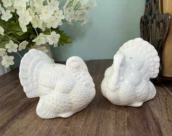 May include: A pair of white ceramic turkey salt and pepper shakers. Each shaker features a detailed molded feather pattern on the body and a fanned tail. The turkeys are depicted in a seated position with heads turned, showcasing a glossy finish.
