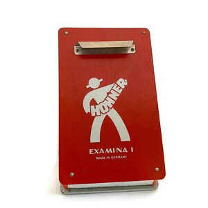 May include: Red rectangular box with a silver metal clip on the top. The box has a white silhouette of a person playing a guitar with the word "HOHNER" written on the guitar. The text "EXAMINA I MADE IN GERMANY" is printed on the box.