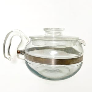 May include: A clear glass teapot with a curved handle and a lid. The teapot has a silver-colored metal band around the middle. The spout is small and curved. The teapot is designed for brewing tea.