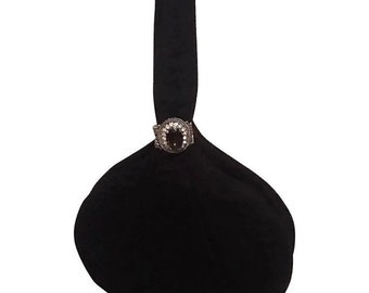 Puede incluir: Un bolso de mano de tela negra con una sola asa superior y un cuerpo redondeado en forma de bolsa. El bolso presenta un broche decorativo ovalado prominente en la base del asa, incrustado con pequeñas piedras claras alrededor de una piedra oscura.