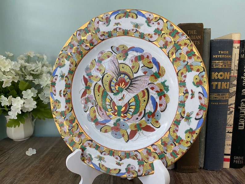 May include: A white porcelain plate with a gold rim and a colourful butterfly design. The butterflies are painted in shades of blue, red, yellow, and green. The plate is on a white stand.