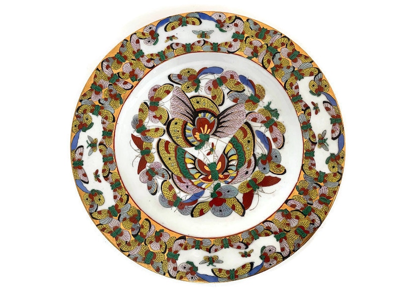 May include: A white ceramic plate featuring a colourful butterfly design. The plate has a yellow border with a repeating pattern of butterflies. The centre of the plate showcases a large butterfly with intricate details in shades of red, blue, green, and yellow.