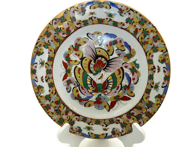 May include: A white porcelain plate with a gold rim and a colourful butterfly design. The butterflies are painted in shades of red, blue, yellow, and green. The plate is decorated with a border of smaller butterflies.
