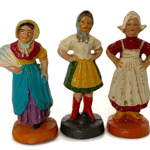 May include: Three vintage figurines of women in traditional attire. The figurines are painted in bright colors, including blue, yellow, red, and green. Each figure stands on a round or square base. One figure holds a fan.