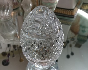 May include: A clear glass egg-shaped container with a diamond pattern on the bottom and a floral design on the top. The container is sitting on a reflective surface.