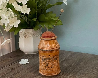 May include: A vintage wooden pin cushion with a cylindrical shape and a small, round, red cushion on top. A long pin with a pearl-like head is inserted into the cushion. The wooden body has a light brown finish with faded text and decorative bands.