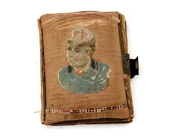 May include: Antique photo album with a portrait of a person wearing a hat and jacket. The album has a brown cover with a decorative border and a black clasp. The portrait is a faded illustration.