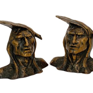 May include: Two bronze busts of Native American figures. Each bust features a detailed face with a stern expression and a feathered headdress. The sculptures have a weathered, antique appearance, with visible texture and variations in color.