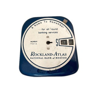 May include: A blue and white vintage bank sign with the text "A Name To Remember... for all 'round banking services" and "Rockland-Atlas National Bank of Boston". The sign has a small, round, white dial with a black hand and a black number display.