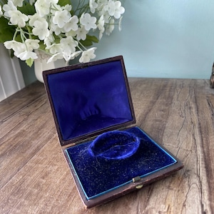 May include: An open, antique jewelry box with a dark brown exterior and a vibrant blue velvet interior. The box is rectangular and has a small clasp. The interior features a circular indentation, likely for a bracelet or ring. White flowers are in the background.