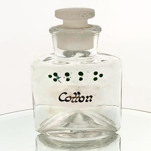 May include: A clear glass bottle with a white lid and a label that says "Cotton". The bottle has green dots around the label.