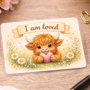 May include: A rectangular card featuring a cartoon Highland cow holding a pink heart. The cow has fluffy orange fur, small horns, and is surrounded by daisies and greenery. The banner above the cow reads "I am loved".