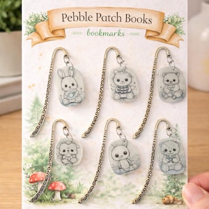 Könnte beinhalten: Eine Sammlung dekorativer Lesezeichen mit niedlichen Tierdesigns, darunter ein Hase, ein Bär und ein Welpe. Jedes Lesezeichen hat einen goldfarbenen, verzierten Metallstiel und einen kieselsteinförmigen Anhänger. Auf der Karte steht "Pebble Patch Books bookmarks."