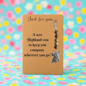 May include: A brown card with the text "Just for you" and "A wee Highland cow to keep you company wherever you go!" A charm with wooden beads and a silver Highland cow pendant hangs from the card.