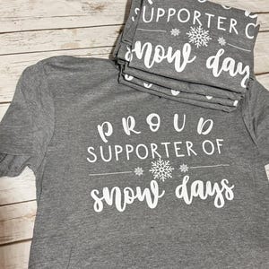 May include: Gray t-shirt with white text that reads "Proud Supporter of Snow Days" with a snowflake graphic.
