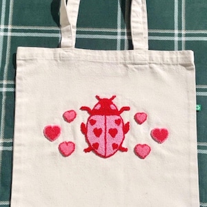 Op de afbeelding: Crèmekleurige draagtas met een rood en roze lieveheersbeestje ontwerp en omringende harten. Het lieveheersbeestje heeft hartvormige details op zijn vleugels. De tas heeft twee handvatten en is gemaakt van canvas.