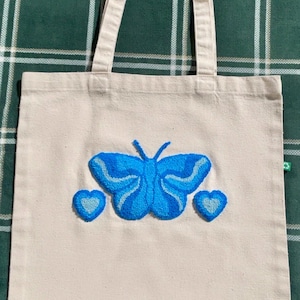 Op de afbeelding: Crèmekleurige draagtas met een blauwe vlinder en twee blauwe harten geborduurd op de voorkant. De vlinder heeft een gedetailleerd ontwerp met verschillende blauwtinten. De tas heeft twee crèmekleurige handvatten.