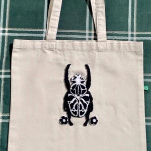 Op de afbeelding: Crèmekleurige draagtas met een zwart-wit geborduurd scarabee-ontwerp. De tas heeft twee handvatten en is gemaakt van stevig canvasmateriaal. Het keverontwerp is gecentreerd op de voorkant van de tas.
