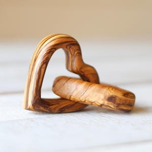 May include: Two interlocking wooden heart shapes, carved from olive wood, with a natural brown and dark brown wood grain pattern.