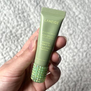 Puede incluir: Un tubo de bálsamo labial Laneige Lip Glowy Balm en color verde salvia, sabor Matcha Bubble Tea. El tubo tiene texto en blanco y una base adornada con pequeños cristales iridiscentes de color verde claro. Peso neto de 10 gramos impreso.