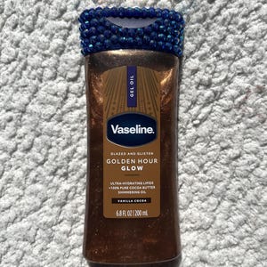Puede incluir: Un frasco de aceite en gel brillante Vaseline Golden Hour Glow con una tapa azul oscuro incrustada de pedrería. La etiqueta dice Glazed and Glisten, Golden Hour Glow, Ultra-Hydrating Lipids, 100% Pure Cocoa Butter, Vanilla Cocoa, 200 ml.