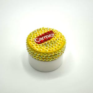 Könnte beinhalten: Ein kleiner, weißer und gelber Behälter mit Carmex-Lippenbalsam. Der gelbe Deckel ist mit schillernden Strasssteinen bedeckt. Das Carmex-Logo ist rot und weiß. Der Behälter befindet sich auf weißem Hintergrund.