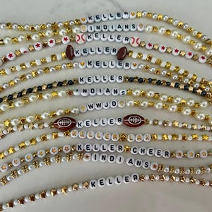 May include: A selection of beaded accessories, including bracelets and mask chains. The designs feature white pearls, gold star-shaped beads, and letter beads spelling out names and words like "KELLER", "INDIANS", and "CHEER". Some have football and baseball charms.