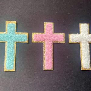 May include: Three felt crosses with gold glitter trim. The crosses are blue, pink, and white.