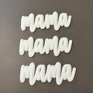 May include: Three white, textured "mama" word cutouts in a cursive font are displayed on a gray surface. The letters have a fluffy, raised appearance, creating a 3D effect. The word is repeated three times.