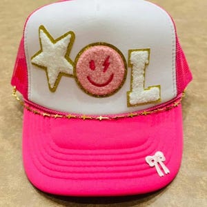 May include: A pink and white trucker hat with a gold chain trim. The hat has a white star, a pink smiley face with a lightning bolt, and the word "LOL" in gold glitter letters.