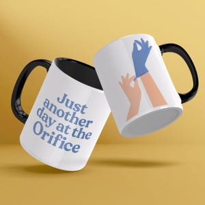 Può includere: Due tazze in ceramica bianca con manici neri. Una tazza ha il testo "Just another day at the Orifice" in lettere blu. L'altra tazza ha un'illustrazione di cartone animato di una mano che si mette un guanto blu.