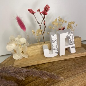 Könnte beinhalten: Weiße Holzbuchstaben, die "JA" buchstabieren, mit einem floralen Design, auf einem Holzregal mit getrockneten Blumen in einer Vase.