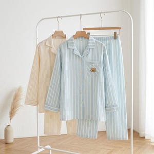 Könnte beinhalten: Zwei Pyjama-Sets hängen an einem weißen Gestell. Ein Set ist hellgelb mit vertikalen Streifen, das andere hellblau mit vertikalen Streifen. Das hellblaue Set hat einen kleinen Bärenaufnäher auf der Brusttasche.