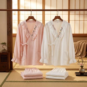 Könnte beinhalten: Zwei passende Pyjama-Sets, eines rosa und eines weißen, hängen an Holzbügeln. Jedes Set besteht aus einem langärmeligen Oberteil mit Rüschenkragen und gestickten floralen Details sowie passenden Shorts. Das Wort "Beautiful" ist auf dem Kragen gestickt.