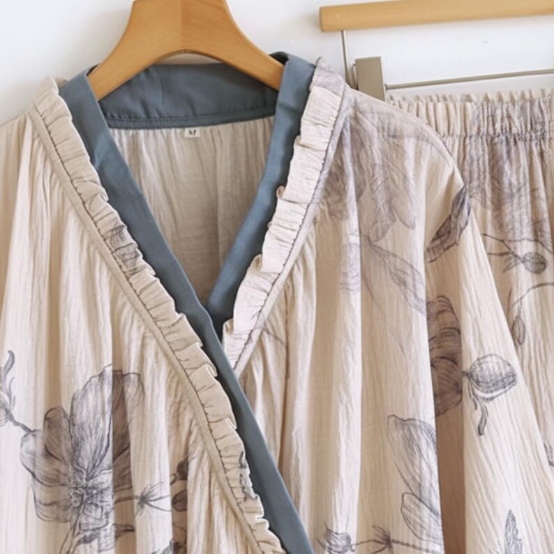 May include: A beige floral pajama set. The top features a V-neck with a blue trim and ruffled edges. The matching trousers are visible on the right. The set is displayed on a wooden hanger.